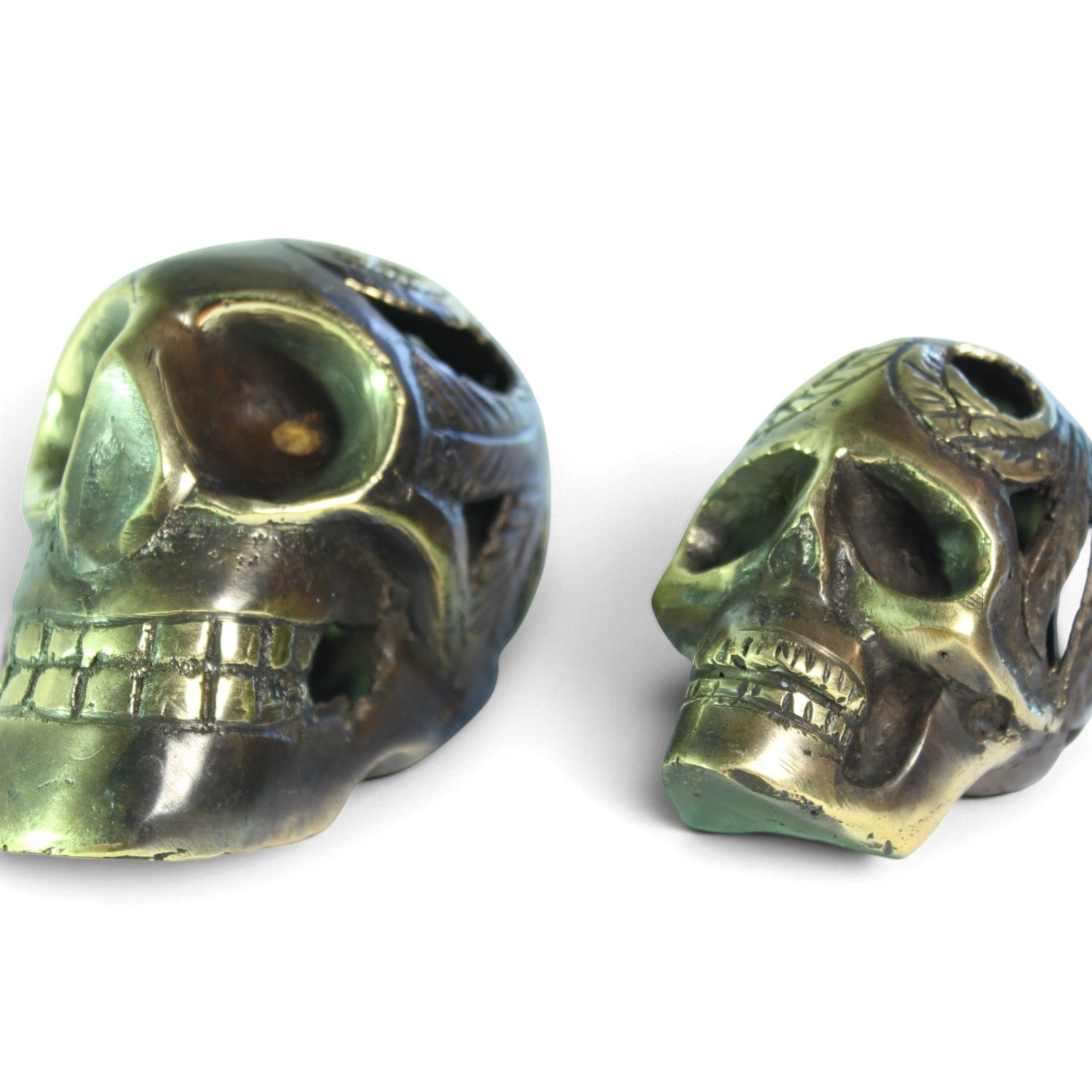 Two brass skull figurines shown side by side, front and profile views, highlighting patina variation